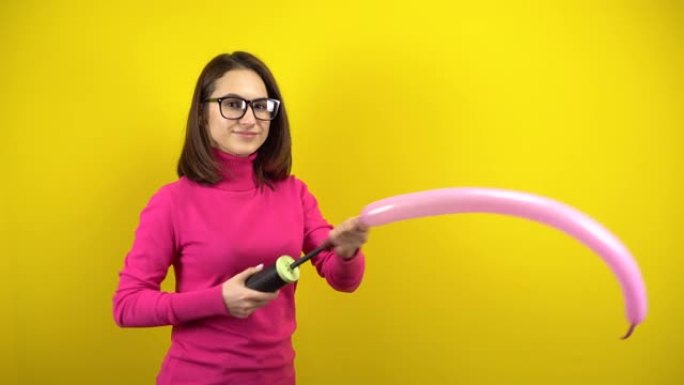 一名年轻女子用黄色背景上的泵给一个长长的粉红色气球充气。穿着粉色高领毛衣和眼镜的女孩。