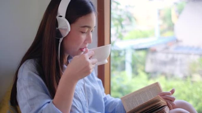 用耳机关闭可爱的女孩读一些书，坐在玻璃窗前，看起来她有快乐的情感