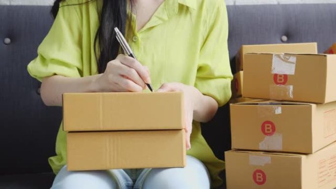 亚洲年轻女性的特写手在家里的包裹箱上写下地址，用于向客户，运输和物流，在线商家和卖家，企业主或中小企