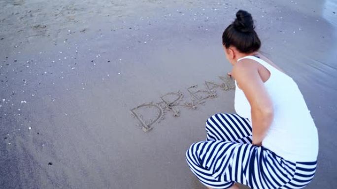 岸上女性的后景。女人在海边的沙滩上写梦这个词。水浪冲走了文字。顶视图。