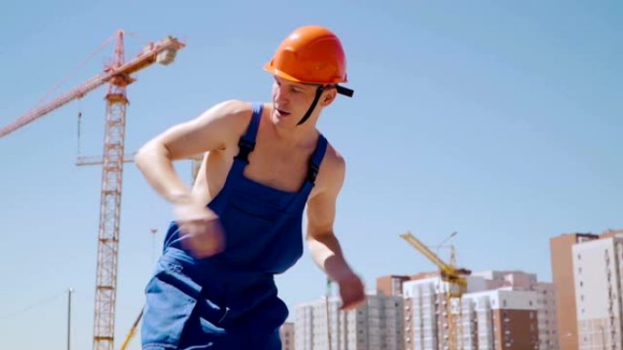 戴着安全帽跳舞的快乐高加索人建设者。工人在建筑工地的背景下滑稽动作。慢动作。斯蒂安卡姆射击。