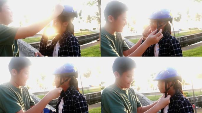 亚洲男孩在早上带着太阳光在公园玩耍之前，为他的妹妹戴着运动头盔。