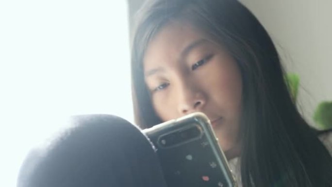 亚洲女孩坐在椅子上，窗灯和使用智能手机在早上在家里，生活方式的概念。