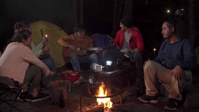 4k亚洲男女坐在篝火旁晚上在森林里享受户外派对。快乐的朋友玩得开心，一起喝啤酒，吃烧烤，一起唱歌。友