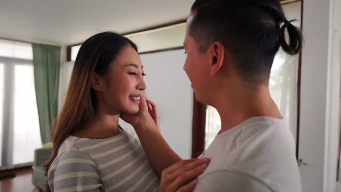 30多岁的亚洲年轻人在家里的客厅里抚摸着一个微笑的女人的脸颊。温柔浪漫的夫妻在舒适的房间里度过家庭时