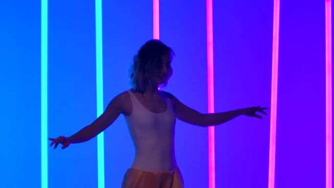 苗条的舞者移动臀部，练习萨尔萨舞的动作。一个女人在慢动作的蓝色和粉红色霓虹灯的背景下在工作室里跳舞。