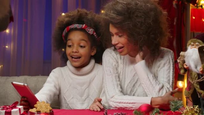 非裔美国妈妈和穿着白色毛衣的小女儿通过平板电脑上的视频通话交谈。快乐全家福，新年庆祝概念。特写