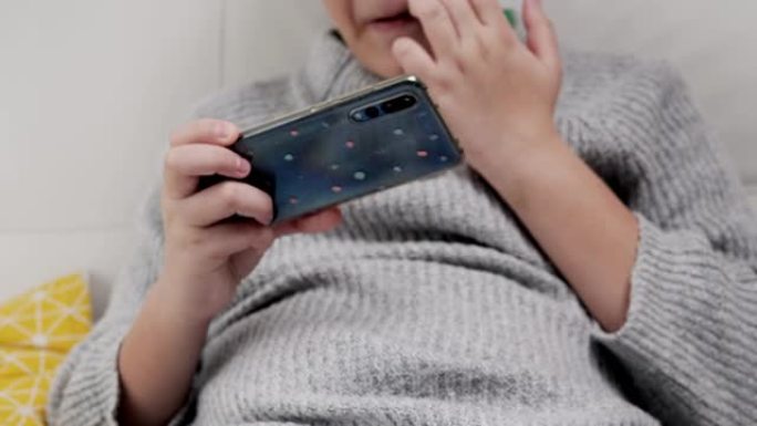 男孩坐在家里的沙发上玩智能手机在线游戏，生活方式概念。