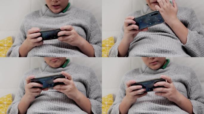 男孩坐在家里的沙发上玩智能手机在线游戏，生活方式概念。