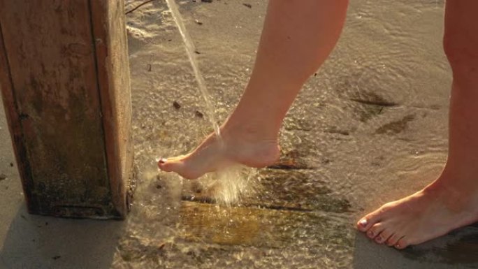年轻女子在沙滩上的沙滩淋浴下从腿上去除沙子，早晨的阳光照在背景中，细节上的水滴落在脚上