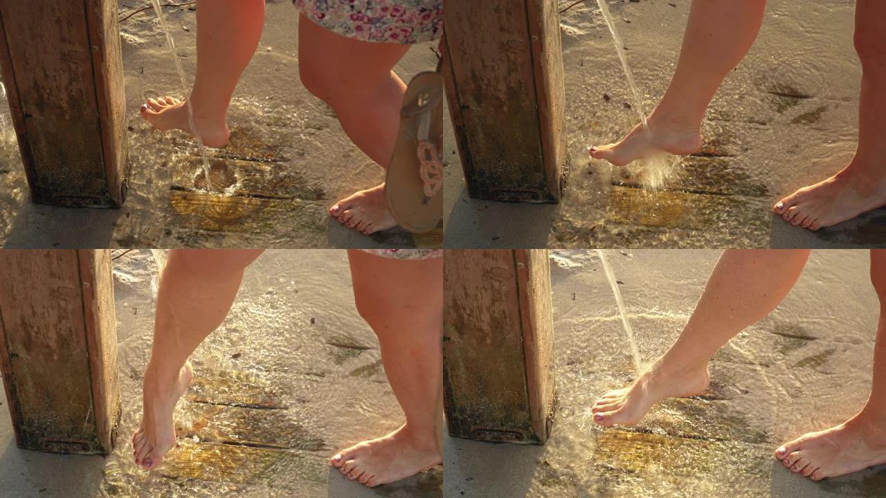 年轻女子在沙滩上的沙滩淋浴下从腿上去除沙子，早晨的阳光照在背景中，细节上的水滴落在脚上
