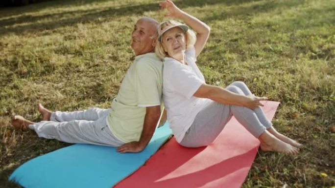 在公园健身锻炼休息时，开朗的老年男女背靠背坐在垫子上并互相交谈的高角度