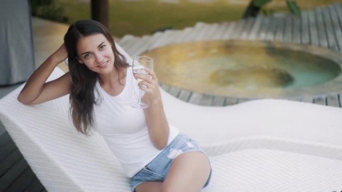 美女坐在游泳池附近的日光浴床上，手里拿着一杯干净的水