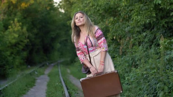 美丽的女人手提箱在夏季森林背景下看着火车上的火车。带着旅行箱的候车女在铁路上偷看晚班火车。