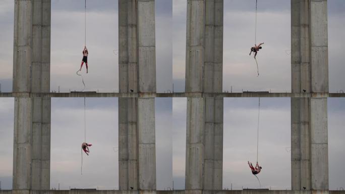 强壮肌肉的马戏团艺术家在空中绳索上表演天空背景上的两个混凝土柱之间的室外，力量、风险和危险的概念