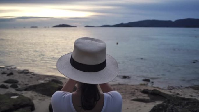 一位穿着白色连衣裙和白色帽子的年轻亚洲女性游客的后视照片正在用智能手机拍摄日落大海的照片