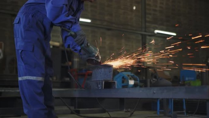 亚洲男技师工人穿着安全的制服耐磨焊工皮革，在工厂车间的钢结构上进行电动轮磨削在车库中，橙色的散景火花