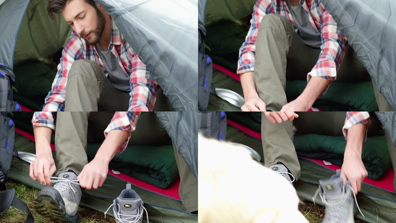 年轻人徒步旅行后系鞋带的特写镜头。美丽的男人和他的狗在帐篷里放松。露营和动物激情