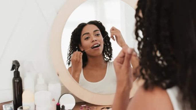 年轻的非洲裔美国妇女坐在镜子前，用牙线在浴室刷牙，后视图