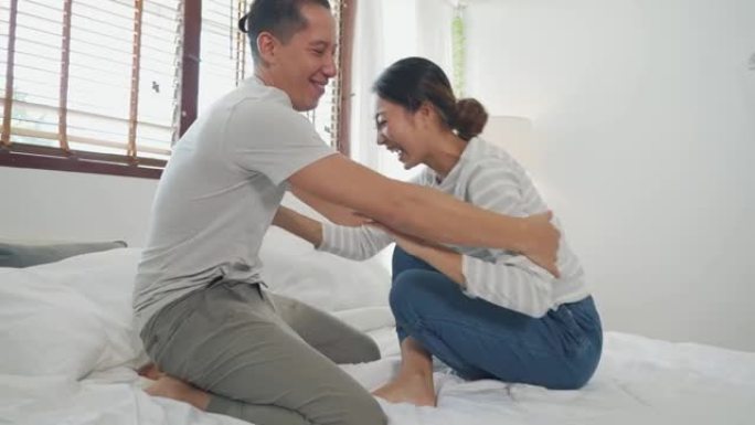在卧室室内场景中，快乐的年轻亚洲夫妇在床上一起挠痒痒的肖像。30多岁坦率成熟的丈夫和妻子微笑。婚姻与