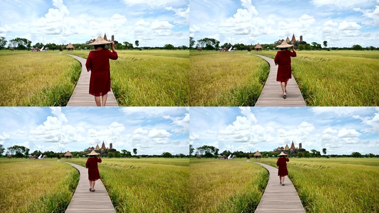 在稻田和Wat Tham Sua视图中，戴着草帽的成年妇女在木路上行走