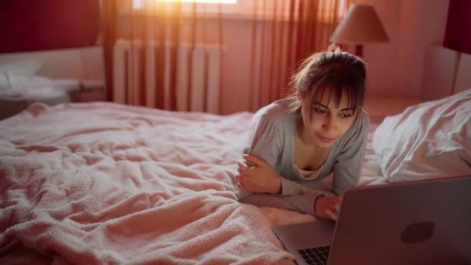 年轻女子躺在家里的床上，看着笔记本电脑，在互联网上浏览