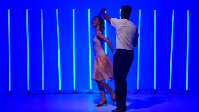 灵活而感性的舞者在黑暗的工作室中，在明亮的蓝色霓虹灯的背景下练习一系列快速旋转。微笑的女孩正牵着她的