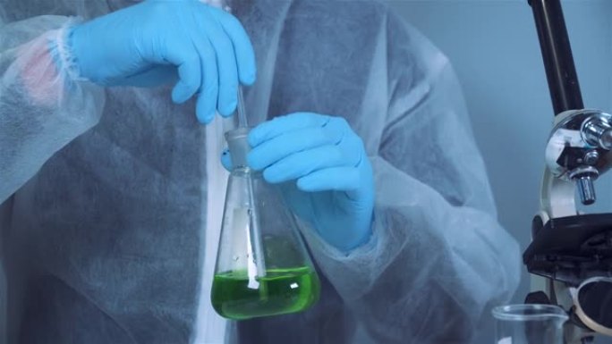 化学实验室防护服技术员在锥形瓶中手动搅拌和摇动绿色液体。实验室技术员研究化学反应并寻找抗癌药物。