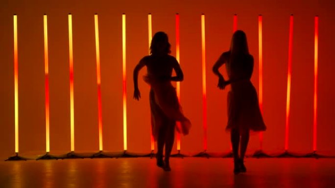 两位优雅的年轻女性在明亮的红色橙色霓虹灯的背景下，在黑暗的工作室里练习萨尔萨舞的元素。轮廓。慢动作