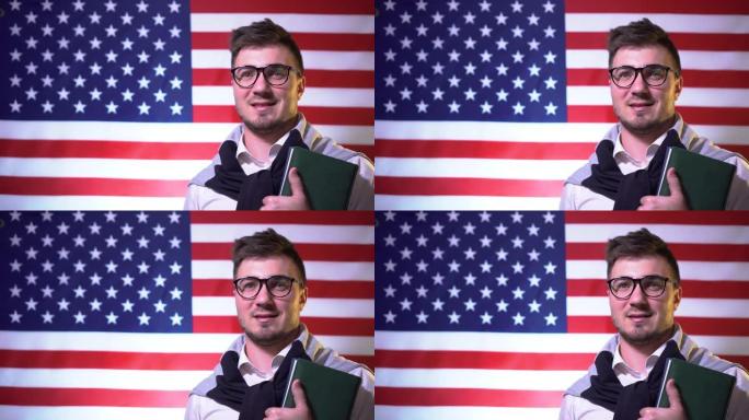 一个戴着眼镜，拿着书，穿着衬衫的漂亮学生站在美国国旗前，微笑着看着镜头。美国的教育。教育理念