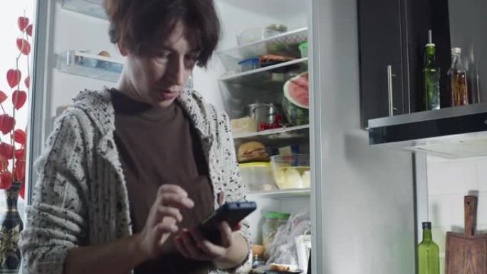 成熟的女人正在打开的冰箱旁边使用手机