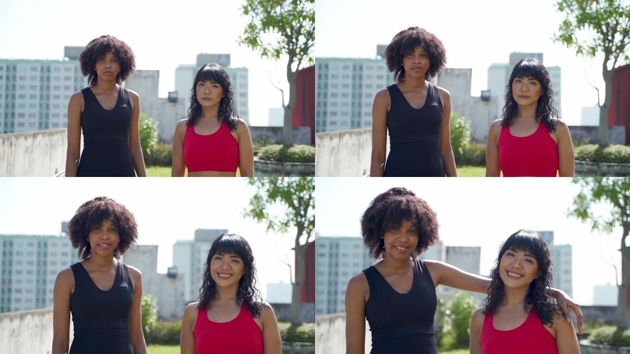成年的两个多民族朋友穿着运动服看着相机，在户外公园微笑。非裔美国人和东南亚妇女站在一起
