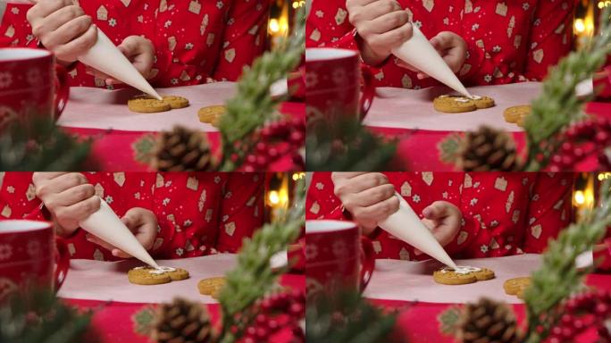 女性的手用糖霜装饰传统的圣诞饼干。穿着红色睡衣的女人，在新年假期装饰的厨房背景上印有节日印花。特写。