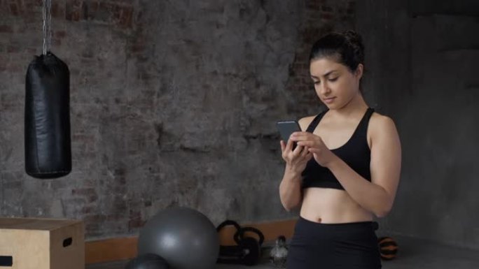 一位穿着黑色绑腿和上衣的年轻印度运动女子使用智能手机观看锻炼程序，并在健身房与教练在线聊天。