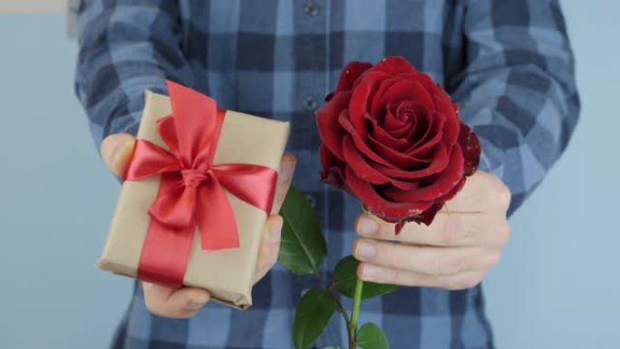 男人正在向相机展示礼物盒和红玫瑰。手拿情人节或国际妇女节的礼物。男送礼盒绑红蝴蝶结，特写