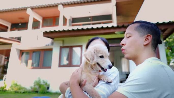 年轻的亚洲成年夫妇抱着一只背景为家庭内部的小狗。30多岁的成熟男人在花园里抱着一只狗宠物。夫妻一起住