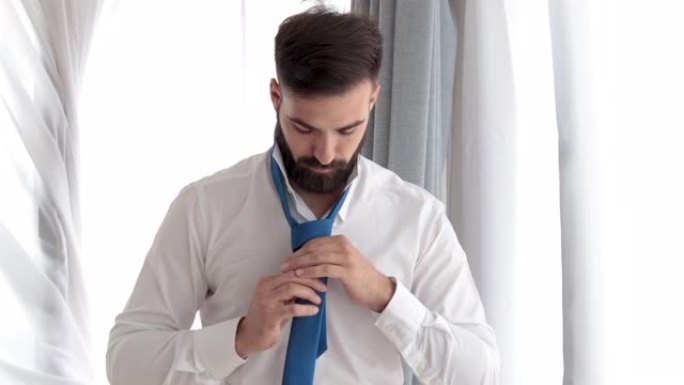 留着胡须的年轻商人男子系好领带，准备在新公司参加新工作面试，或者他是准备婚礼的新郎