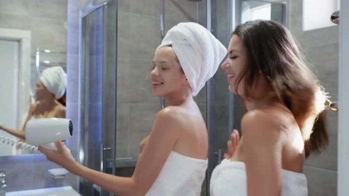 生活方式，穿着毛巾的迷人年轻女性在淋浴时玩得很开心，对着吹风机唱歌，就像对着麦克风在浴室里笑着唱歌