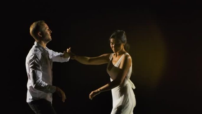 可爱的微笑夫妇跳舞萨尔萨在一个黑暗的工作室。一个穿着白色衣服的男人和一个女人正在欣赏舞蹈。缓慢的运动