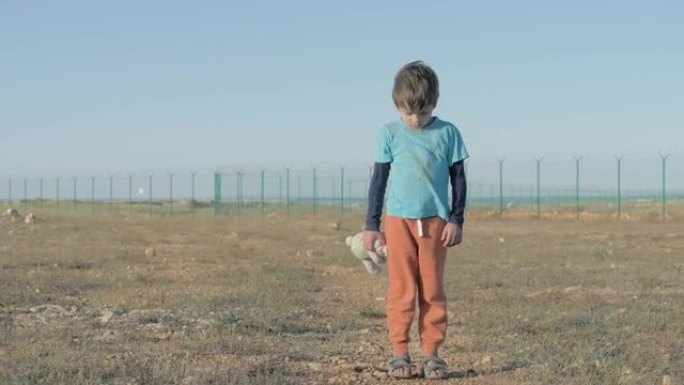 穿着脏衣服的难民小孩。难民营被高高的围栏包围着，可怜的小男孩抱着毛绒兔子望向远方