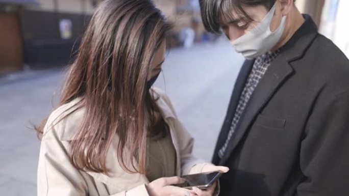 在大风天，在街上使用智能手机戴着防护口罩的年轻夫妇