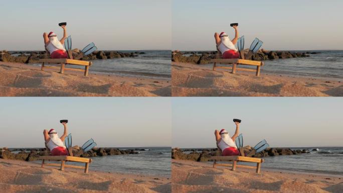 圣诞老人日光浴。戴着墨镜和脚蹼的有趣的圣诞老人坐在躺椅上，在海边的海滩上，手持小型音乐扬声器听音乐，