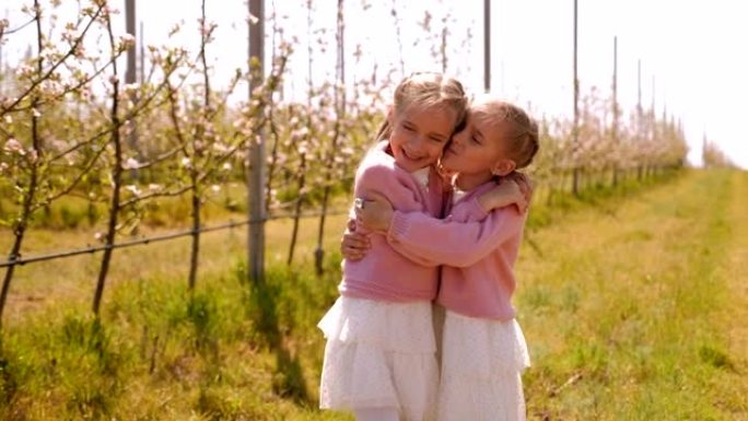 春天，两个穿着相同衣服的双胞胎姐妹在苹果园拥抱。
