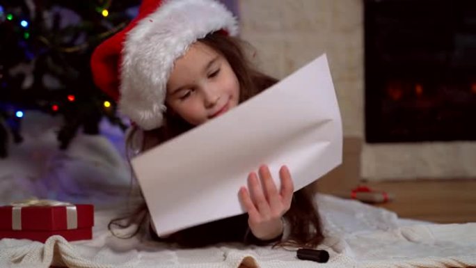 戴着圣诞老人帽子的微笑女孩写信给圣诞老人。圣诞奇迹愿望清单。孩子躺在带圣诞灯的圣诞树前装饰好的家里。