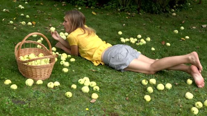 穿着黄色衬衫的美丽女性躺在草坪上，在柳条筐附近用水果吃苹果。4K