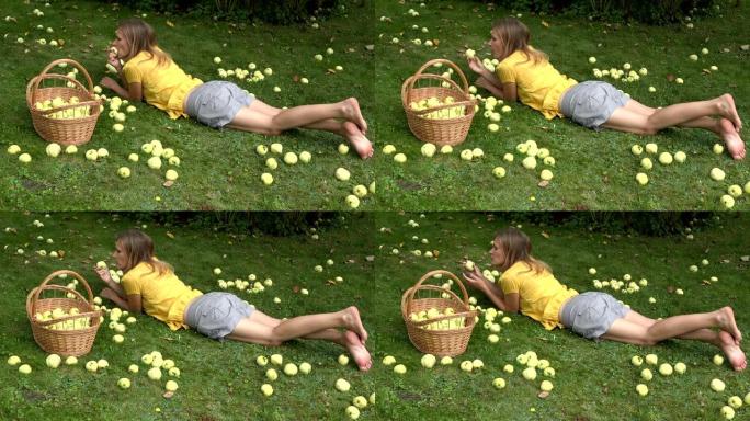 穿着黄色衬衫的美丽女性躺在草坪上，在柳条筐附近用水果吃苹果。4K