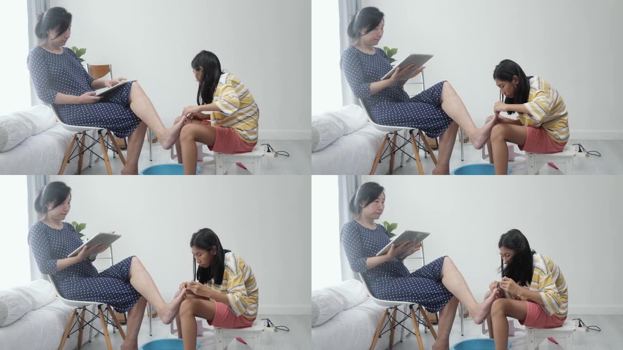 快乐的亚洲母亲使用数字平板电脑，而女儿在脚趾甲绘画前清洁脚，生活方式概念。