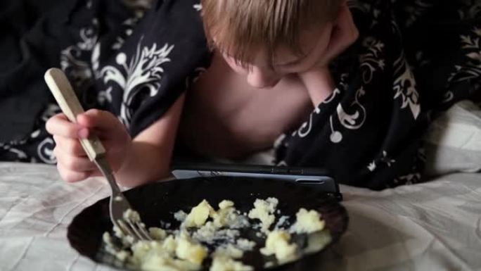 一个小男孩正在床上吃饭，看着手机。