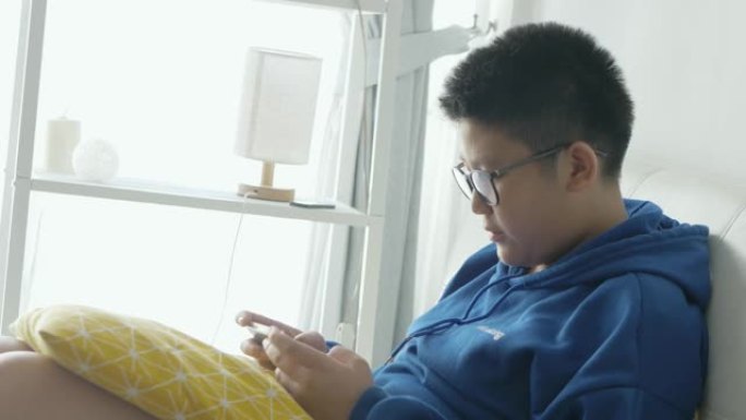 亚洲男孩戴着眼镜，坐在家里的沙发上，通过智能手机玩网络游戏，生活方式概念。