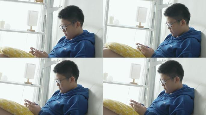 亚洲男孩戴着眼镜，坐在家里的沙发上，通过智能手机玩网络游戏，生活方式概念。